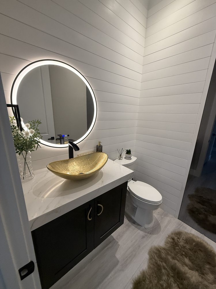 Powder room with white horizontal wall paneling, black vanity, gold vessel sink and round illuminated mirror