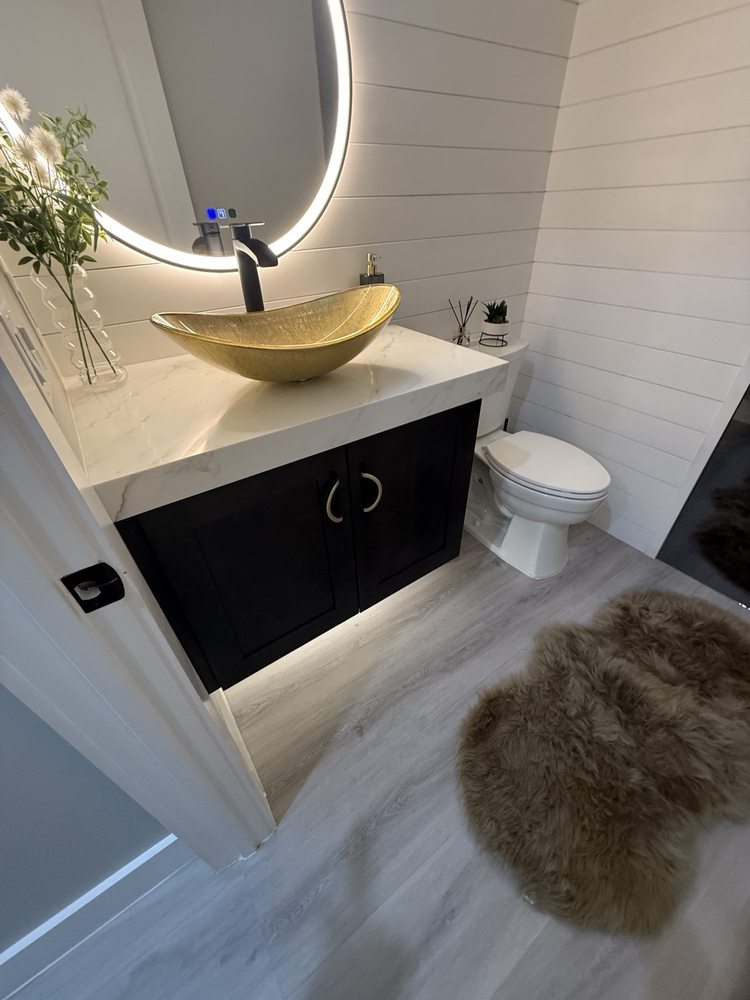 Black vanity, gold vessel sink, white countertop and round illuminated mirror